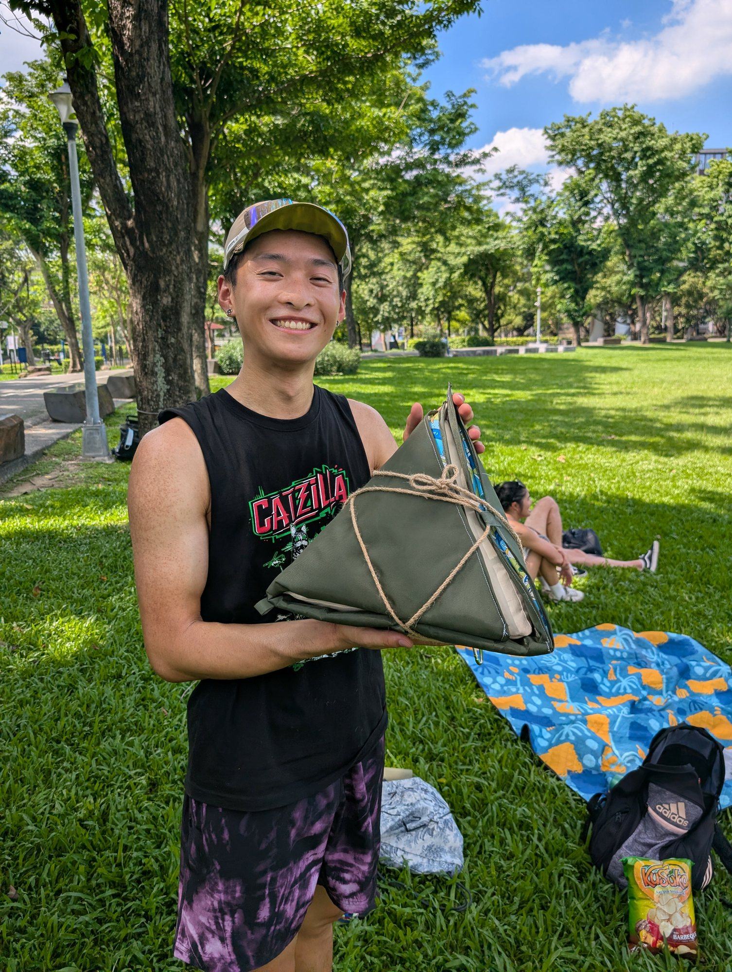 Jason standing in a park holding the Zong Zi wrapped into a compact olive triangle bundle tied with cord.